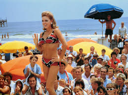 A tanned woman in a Confederate bikini stands before an appreciative beach crowd.