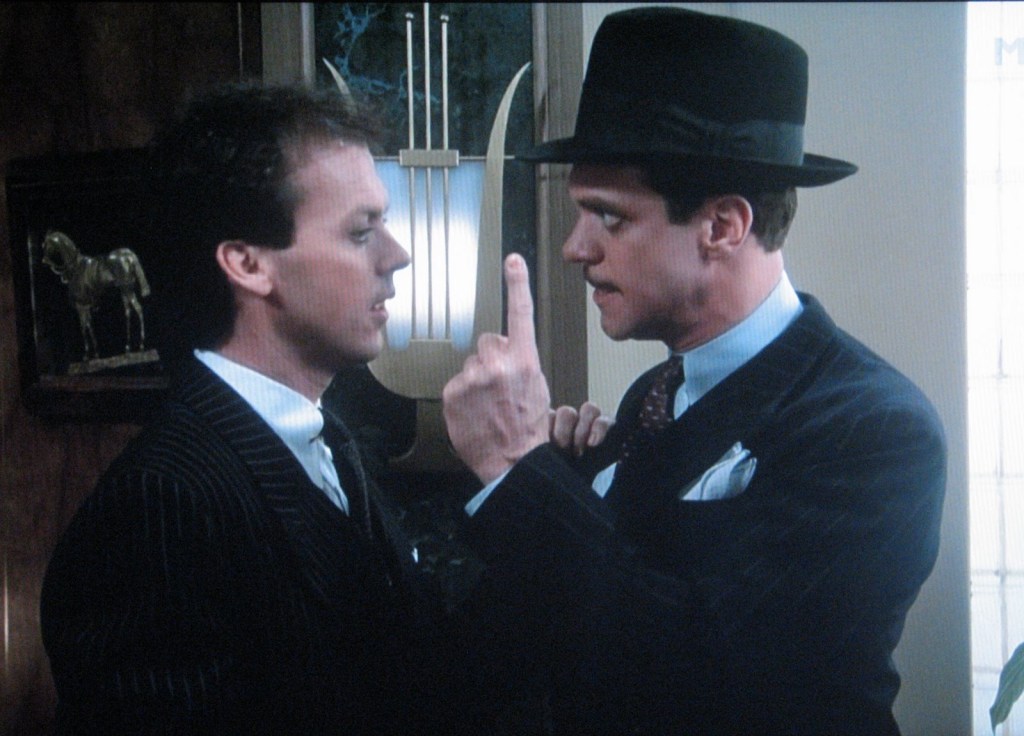 A man in a black suit and rounded black hat holds a finger up to Michael Keaton's face.