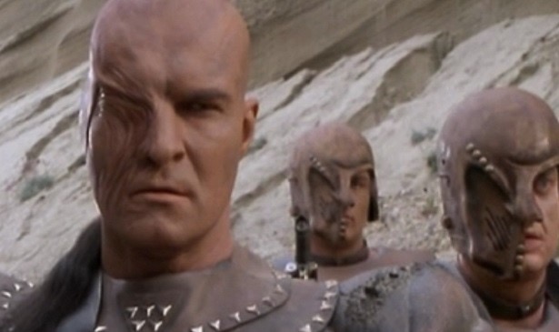 I haven't seen it, so bear with me here. Richard Moll, an imposing bald man, has half of his face scarred in wavy vertical lines. Next to him are two men possibly wear helmets that make them look scarred as well. They all wear tan tops, but Moll's is studded.