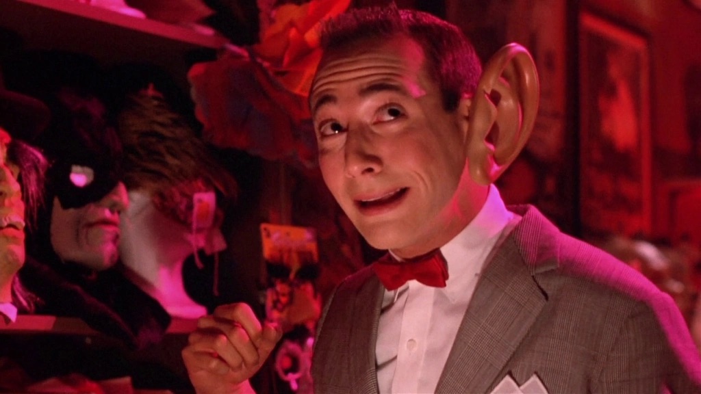 Pee-Wee, a thin, childlike man in a gray suit, in a costume store trying on an oversized ear.