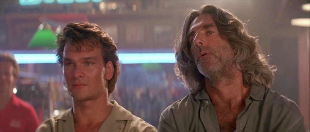 Two white men, Patrick Swayze and Sam Elliot with fabulous long hair, stare at something the camera doesn't see.