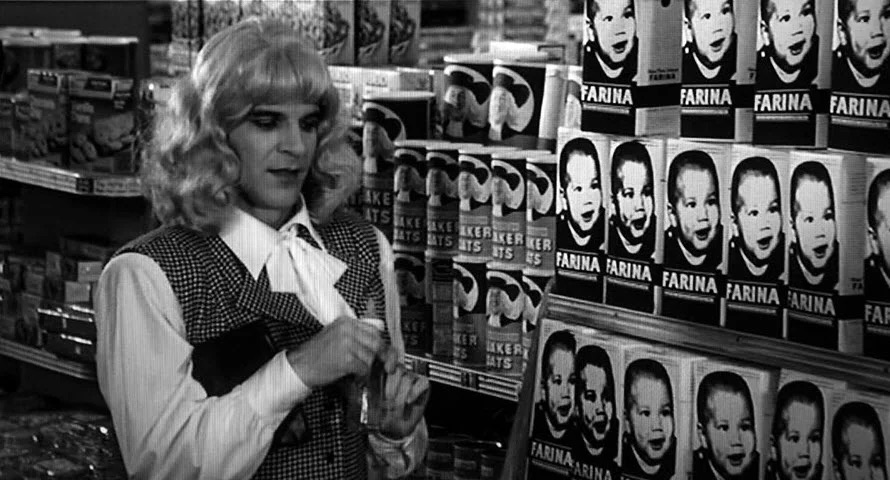 Black and white, Steve Martin in a wig and dress stands in front of a grocery aisle with only two items filling the shelves, oats and something that's probably like oats and doesn't exist anymore.