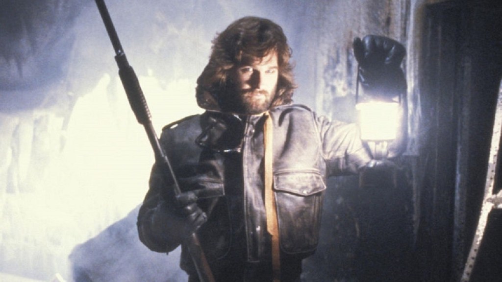 A bundled-up Kurt Russell holds up a lantern while his other hand holds a long thin gun, may e.