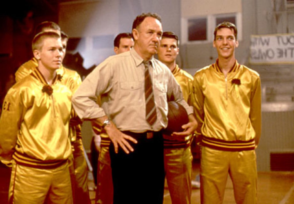 Gene Hackman stands seriously with hands on hips, a basketball tucked into one arm, with young, yellow-clad athletes standing around him.