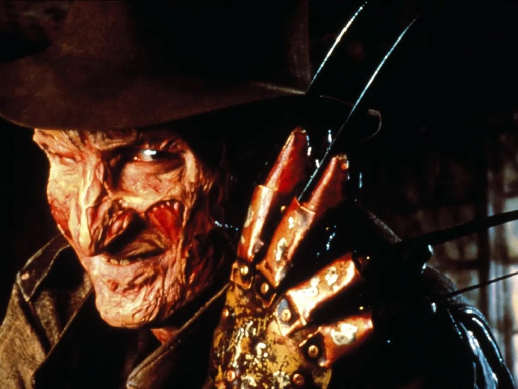 A close up of Freddy, a smiling burned man wearing a hat, with his knived metal glove up next to his face