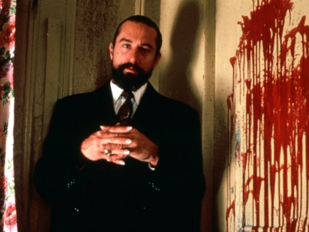 A bearded man in a suit folds his hands and looks forward. Next to him is a red-splattered wall. Creepy!