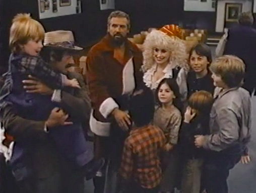 Dolly Parton in what might be a very Parton-esque elf costume–ruffled white shirt, black vest, cleavage, possibly a knitted cap barely resting on her curls but the picture is fuzzy–with a man in a Santa shirt and another holding a child. Children surround them.