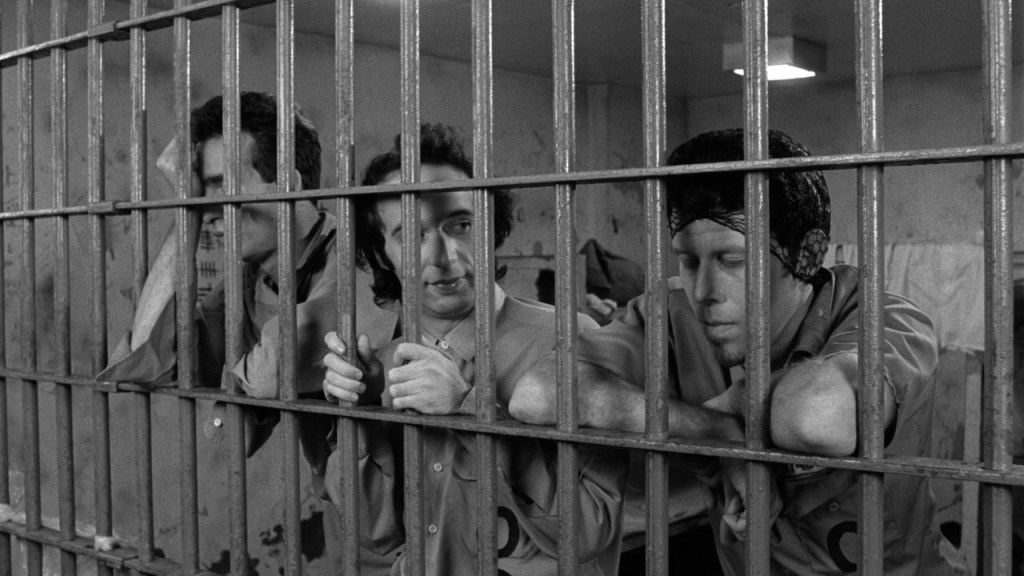 Black and white. Three men behind bars lean against the cell door.