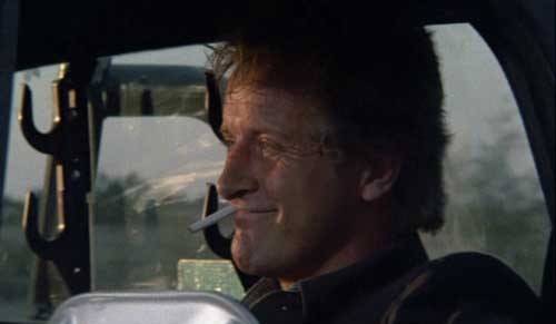 Rutger Hauer in a car smoking a cigarette and smiling.