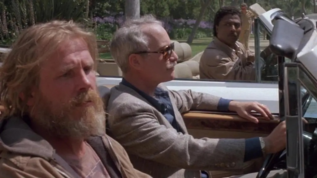 A Black man looking startled at the car next to him, which holds a white man driving and another that is obviously down and out.