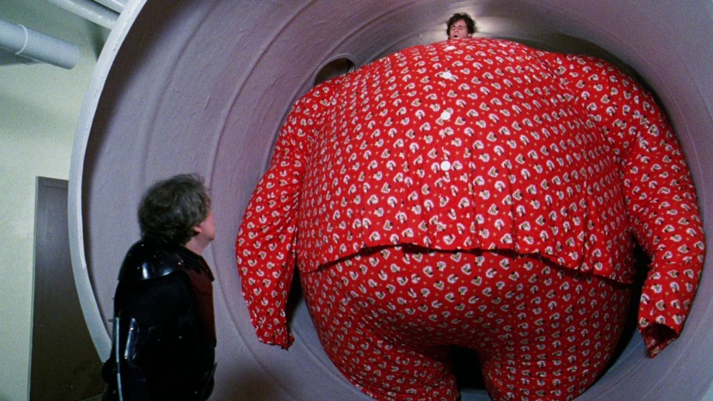 A man looks on as another...okay I'm trying here. Rick Moranis's tiny head is on a comically inflated body, and it's very Willy Wonky.