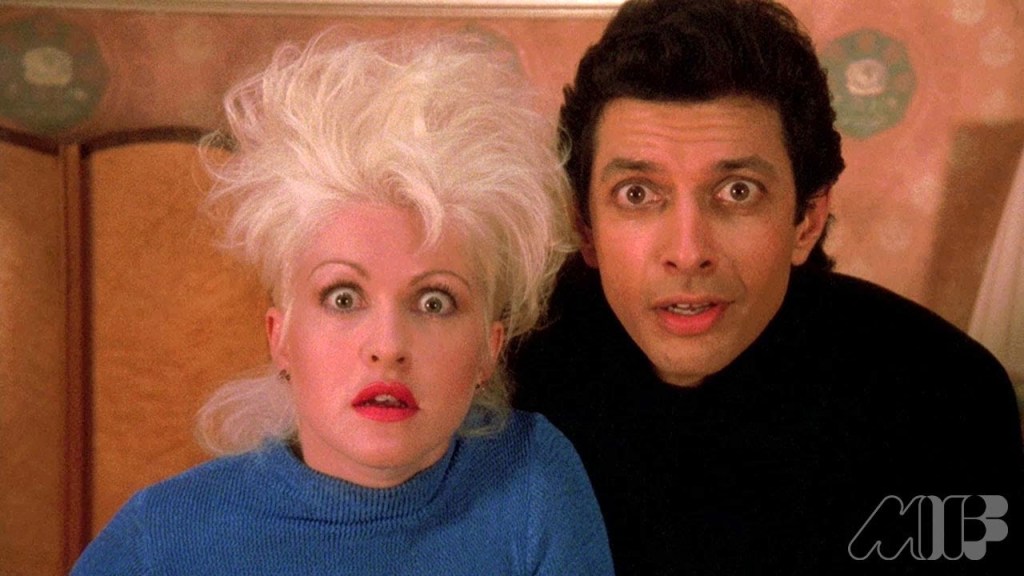 Cyndi Lauper and Jeff Golblum look incredulously directly into camera. She looks so good here. Her bleached hair, pink lipstick, and blue top really pop together.