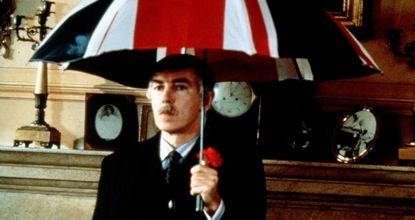 A white man in a black suit and neat mustache is indoors but holds an open umbrella in the British flag colors.