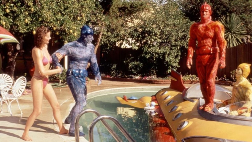 A fuzzy blue alien man leads a bikini'd Geena Davis to a backyard pool, in which two other brightly-colored aliens are hanging out