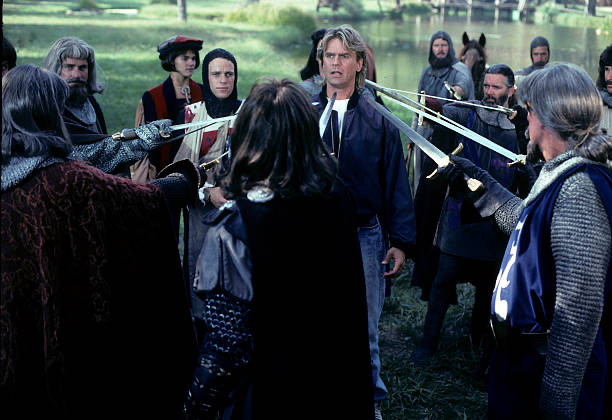A man in jeans, windbreaker, and sort of a mullet shag thing is surrounded by a bunch of knights in terrible wigs, all pointing swords at him.