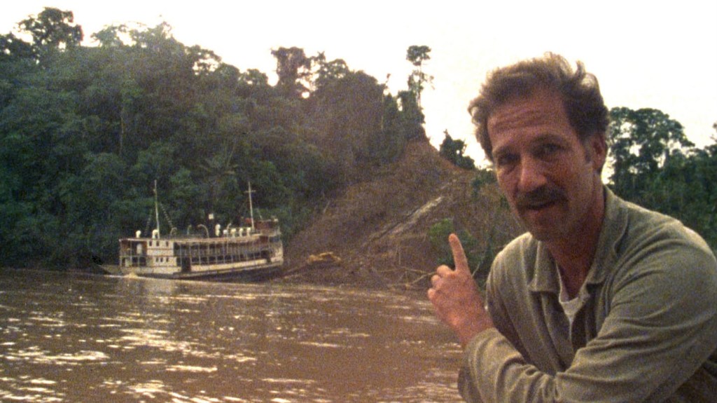 A mustached man points to a boat in the background, which is half on water, half on land.