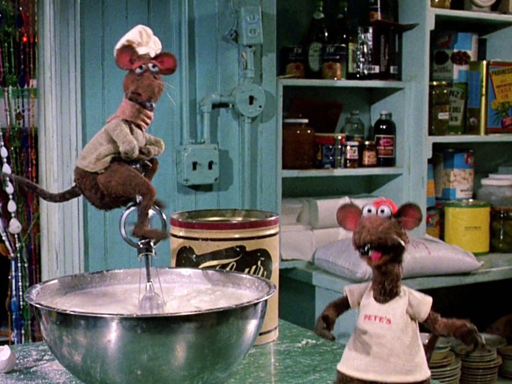 Two puppet rats in a kitchen. One is wearing a white t-shirt that says Pete's as the other wears a chef's hat and rides an egg beater like a unicycle