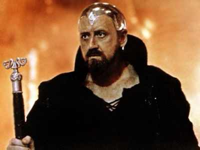 A man in black with a metal plate on his head, which made me think this wasn't the photo for Excalibur. He looks like he's wearing the 80s idea of shiny silver everything and also he might have a computer attached to his brain. but I guess not. He holds a staff and I wonder if he's evil, because he has traditional evil guy facial hair.