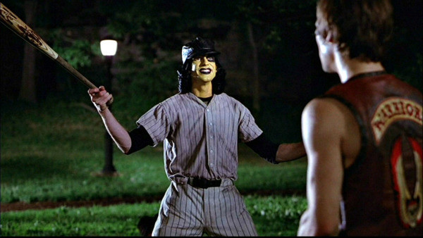 A guy in a baseball uniform with full yellowy neck and face paint waves a bat manically.