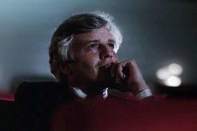 A man with gray in his temples watches an off-camera movie screen in a dark theater. There are tears in his eyes.