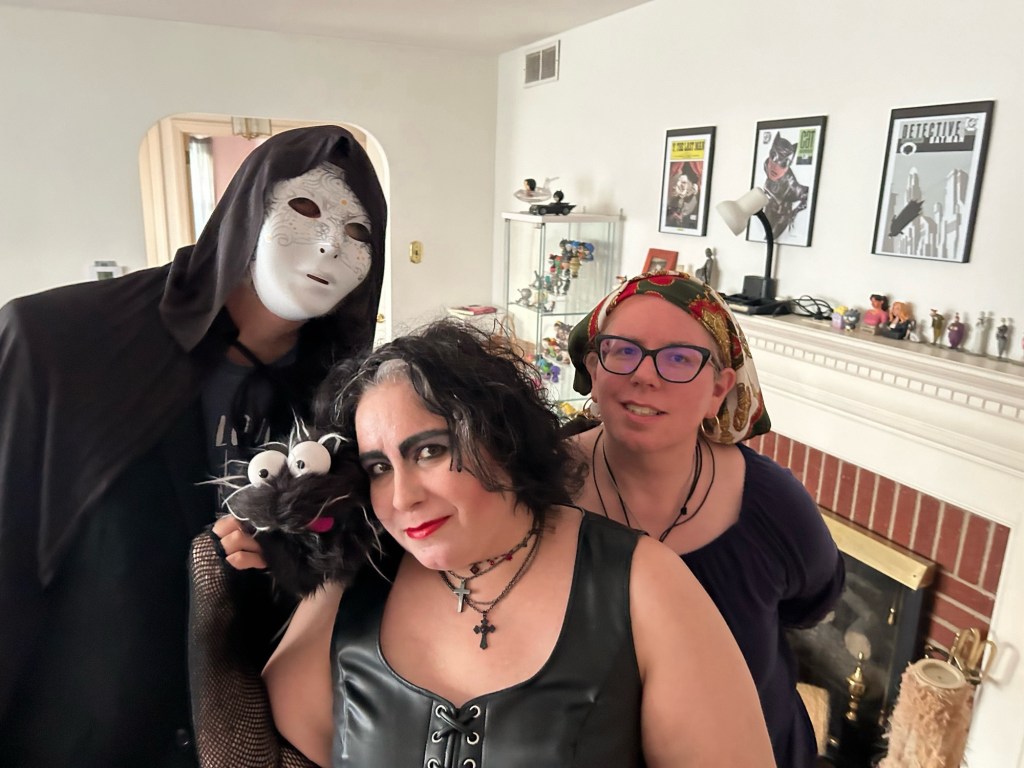 Mary, Dennis, Alana, and Reggie ready for Halloween. Mary wears a headscarf, gold hoops, and a peasant top. Alana (me) is gothed out with Siouxsie Sioux-style eye makeup and her hair dyed black but for one silver streak. She's wearing a pleather top and fishnet gloves, one of which caresses Reggie's little puppet face. Dennis wears a black cape and a creepy white mask that I should probably know the origin of. It's not Ghostface, it's more Eyes Without a Face.