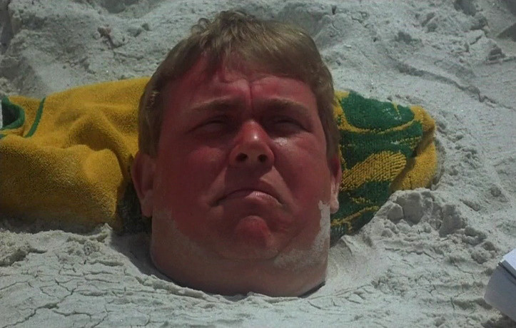 A very sunburned John Candy is buried up to his neck in sand. at least there's a towel behind his head like a pillow?