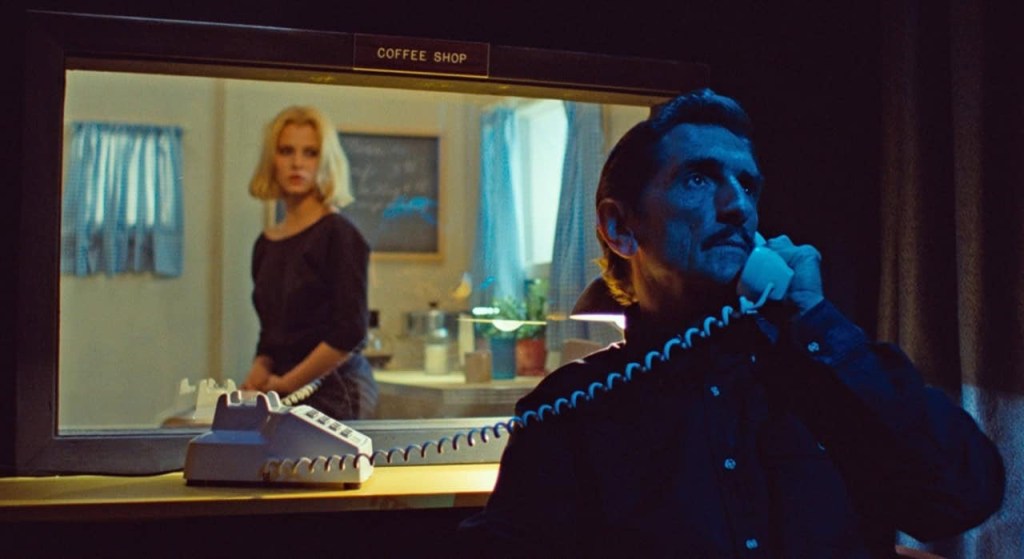 Harry Dean Stanton, hair slicked back and in a button-down shirt, is lit in blue as he speaks on the phone. Behind him in a window is a normally-lit blonde woman in a coffee shop. I know this because above the window on Stanton's side is a little sign that says Coffee Shop.