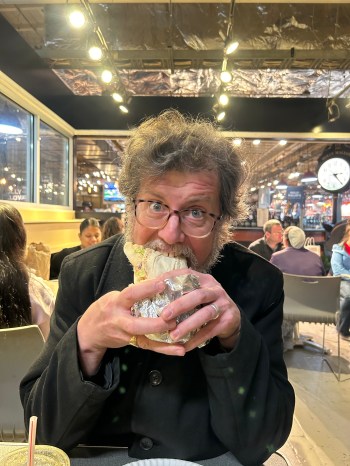 Dennis, a hotter Kurt Vonnegut, is taking a bite of a large burrito. He looks surprised at the picture being taken.
