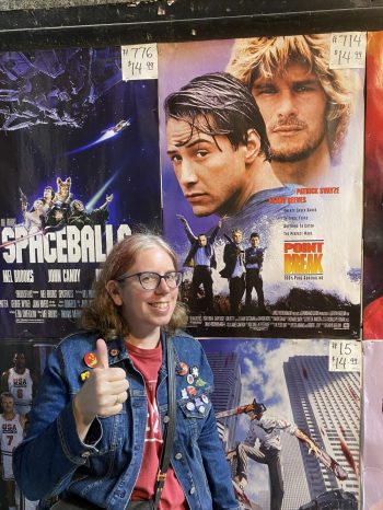 Mary, a white woman with glasses, gray in the part of her otherwise reddish hair, and a great smile, wears a jean jacket studded with buttons and gives a thumbs-up in front of Spaceballs and Point Break movie posters down the shore.
