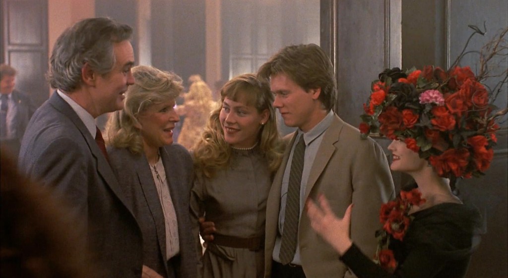 Kevin Bacon and a young woman are talking to an older couple. Jennifer Jason Leigh is there wearing what can only be described as the most extra headpiece I have ever seen. It's like a cross between a Frida Kahlo flower crown, a Goth bush, and an 18th century wig. It's black with red flowers and has a bird in wired branches above like it's in a tree.