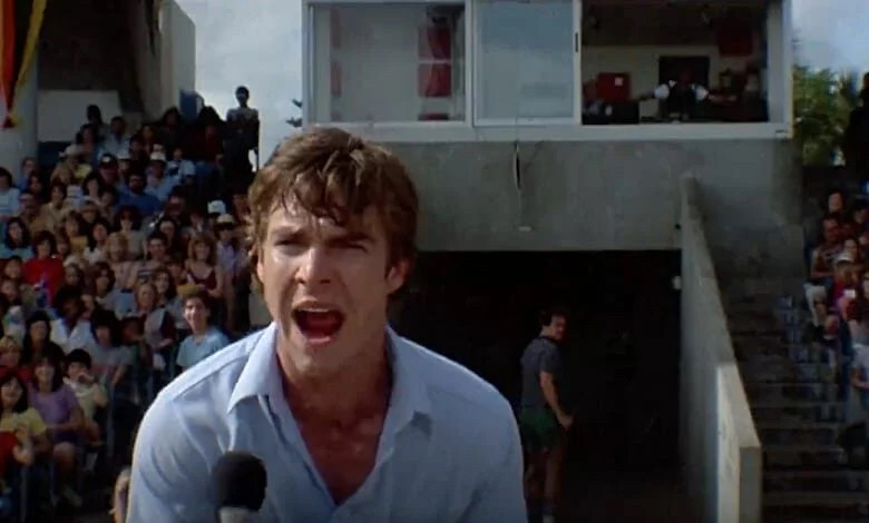 Dennis Quaid screams into a microphone with bleacher-style seating behind him.