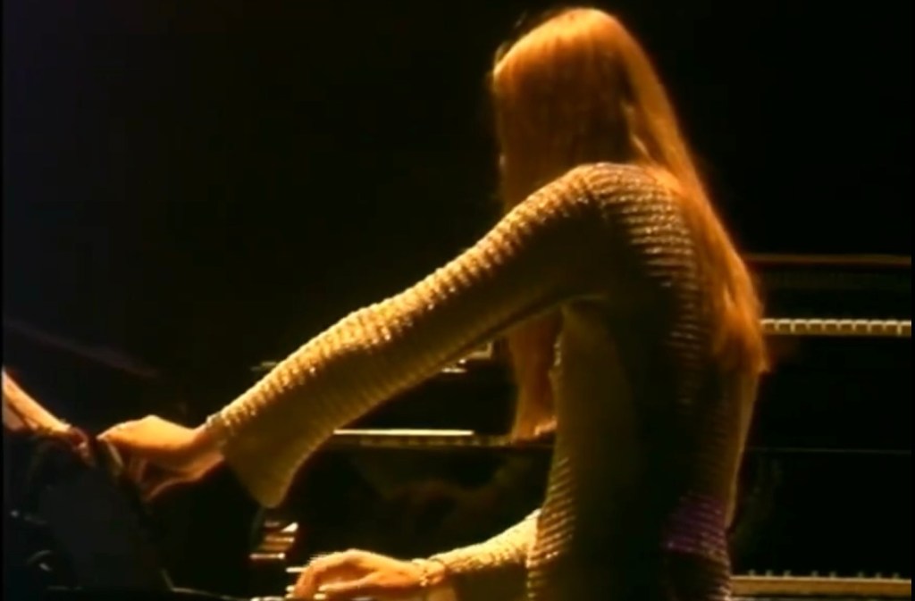 A person with long golden hair faces a keyboard or something like it. I want to make a Tori Amos joke because she, too, would wear a top that might be glittery or might be chain mail.