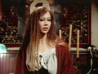 Woman with crimped hair stands in front of a rug on a wall. It doesn't even look like a tapestry. It looks like a rug. It looks like the 80s.
