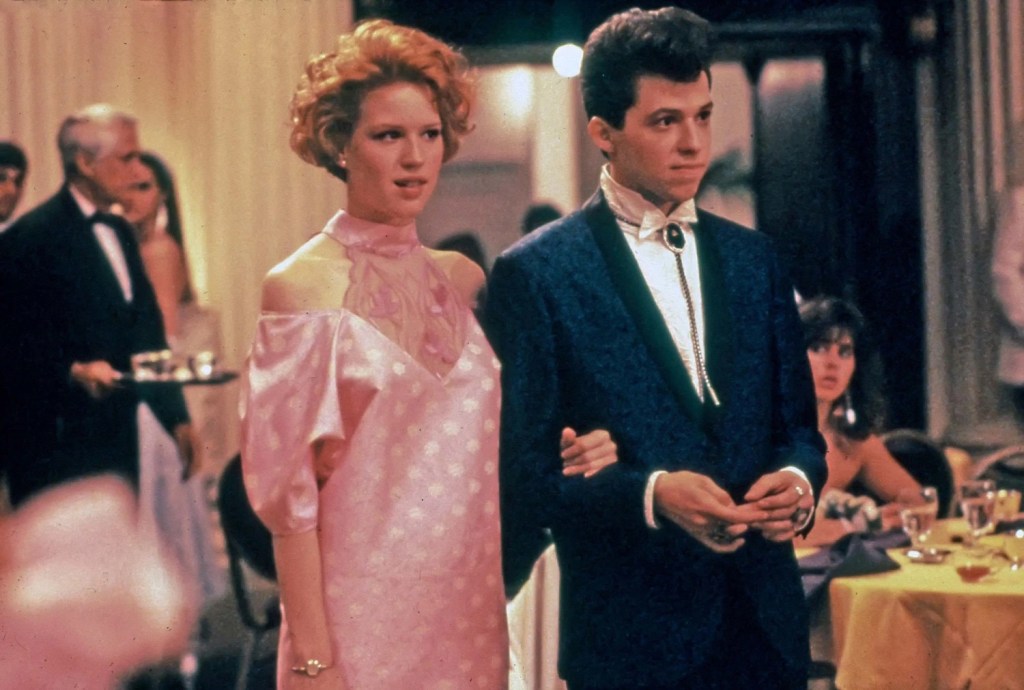 A young redhead in a pink dress is on the arm of a young man who seems to style himself like Lyle Lovett, all big hair and bolo tie. The 80s were wild.
