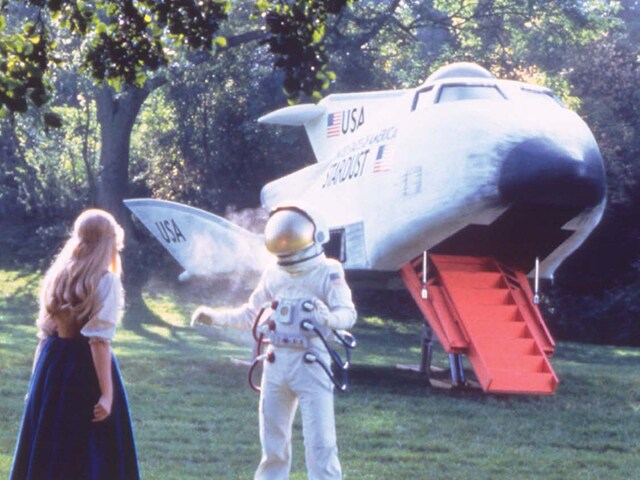 A woman with long blonde hair and her back to the camera looks at a person in a spacesuit with a rocket behind them.
