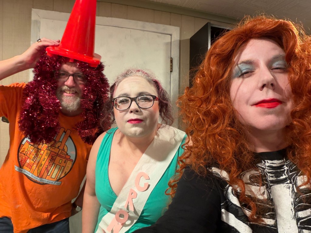 Dennis is dressed as Chappell Cone, with red tinsel hair and an orange traffic cone on his head; Alana is Chappell Crone, wrinkles emhasized with makeup, dressed in tiara and gown and toilet paper sash that says Crone, leaning toward the camera like she didn't quite hear what you said; Mary is Chappell Bone, in a red wig and skeleton sweatsuit.