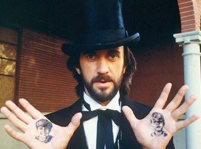 Dennis's alt text from Bluesky: Jonathan Pryce holds up his hands to reveal tattoos of the two boys he’s chasing, Jazz hands style.