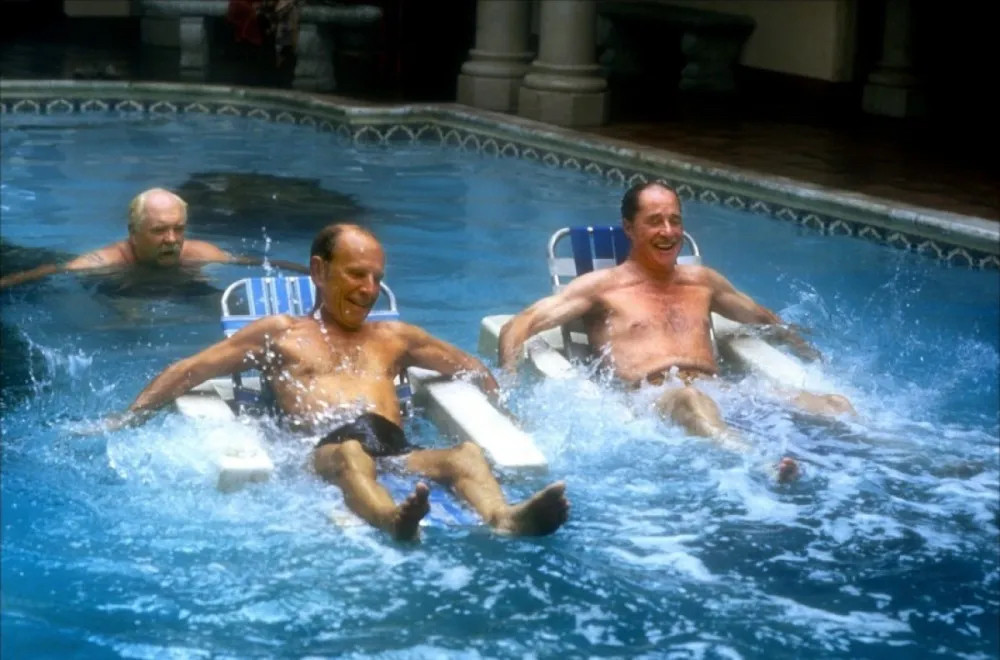 Three old men in a swimming pool–two on those floating chairs and one swimming behind who is Wilford Brimley.