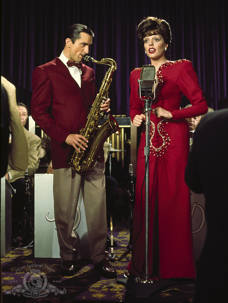 A man with a saxophone and a woman singing at a microphone. His red velvety suit coat sort of matches her dress, though it's a bit darker. There's a band at podiums in the background. It could not be more 1940s.