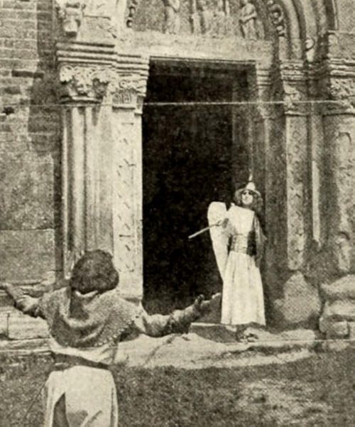 An...angel? points a sword at a knight with a bowl haircut. We only see the back of the knight. They are maybe standing in front of a temple of some sort. it looks like there's a clothesline across the top, but that can't be right.