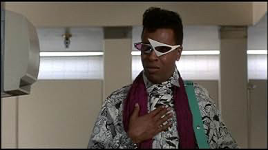 Meshach the God as Hollywood Montrose, in a black and white patterned top, possibly multiple scarves, and his amazing asymmetrical sunglasses, with one big chunk white triangle frame and the other elongated and patterned. Icon.
