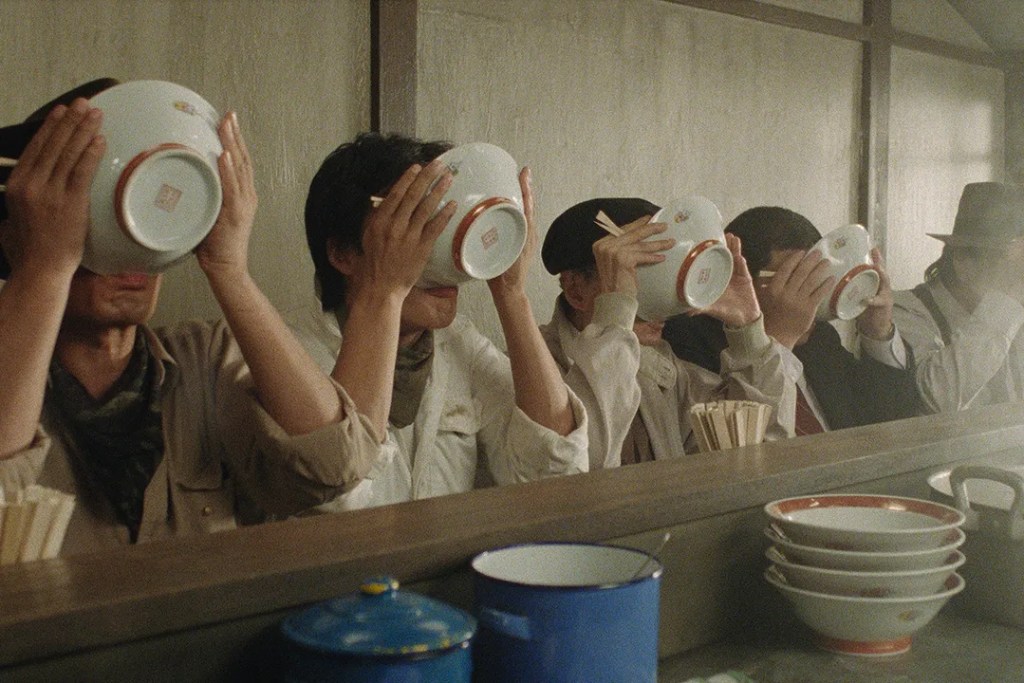 A row of people at a counter, bowls covering their faces to get the last of their meal