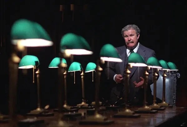 Ned Beatty, looking very old with gray hair, mustache, and a suit, stands in front of a lot of green desklamps