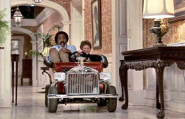 Richard Pryor looks terrified as a gleeful child drives them around in a child-sized car inside an elegant home