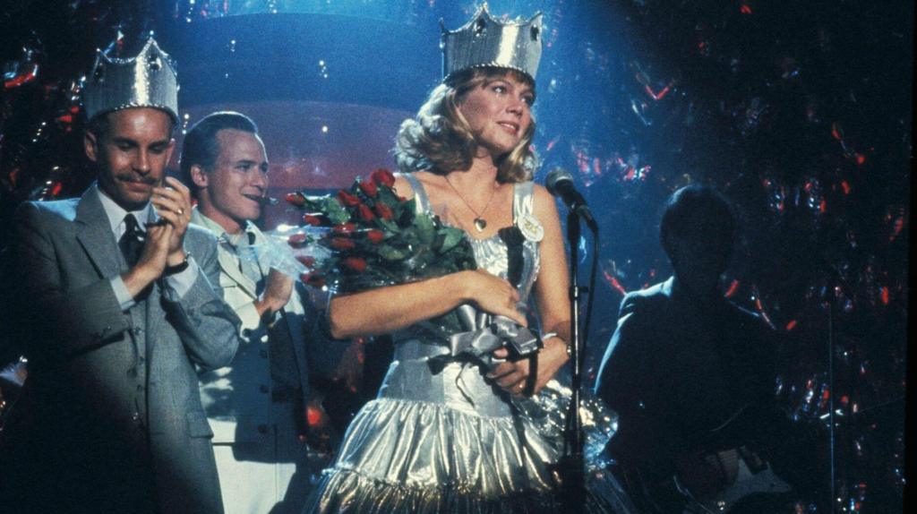 A spotlighted blonde woman in a silver dress wears a silver crown and and armful of flowers. The man next to her is clapping and also is wearing a crown.