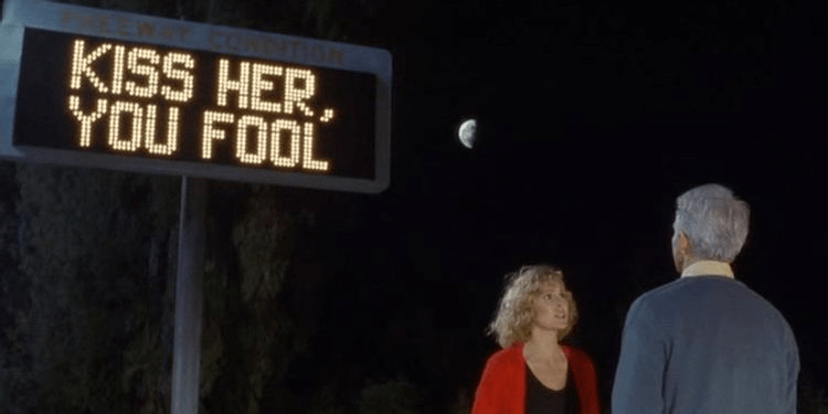 Two people stand beneath a billboard at night. The sign reads Kiss her, you fool.