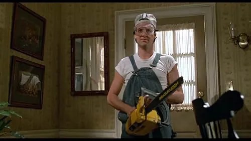 Michael Keaton stands inside a home in a white t-shirt, overalls, safety goggles, and a backwards baseball cap. He is holding a chainsaw.