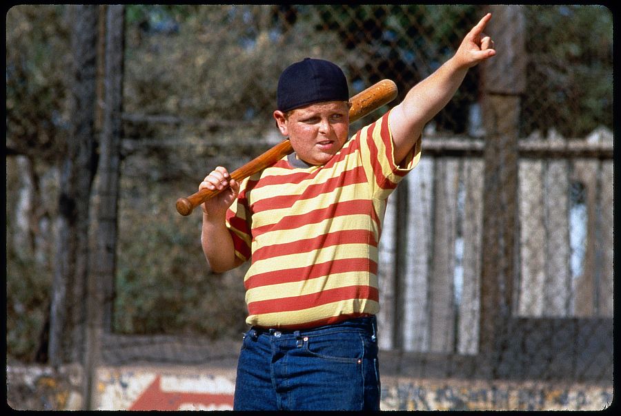 A young white boy with freckles covering his face, a red and white striped shirt, a dark blue backwards baseball cap, and a bat slung over his shoulder raises his arm and points