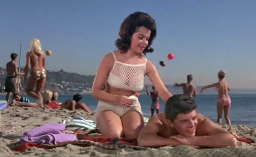 A brunette with big hair is wearing a biking that's got white fishnet on it and I cannot figure out if I love it or I think it looks like a laundry bag. Oh, and she is putting sunscreen or something similar on the back of a man lying on a towel on the beach.