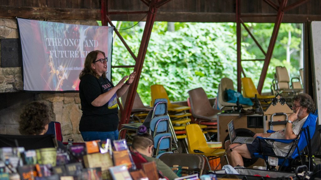 Mary, a white woman in a black t-shirt, holding her hands up and mouth open to speak, standing in front of a screen. She is in what looks like an open structure in the woods. Dennis is in the front of the audience with a laptop open. The screen has the same image as what's behind Mary, so I guess he clicked on the slides for her.